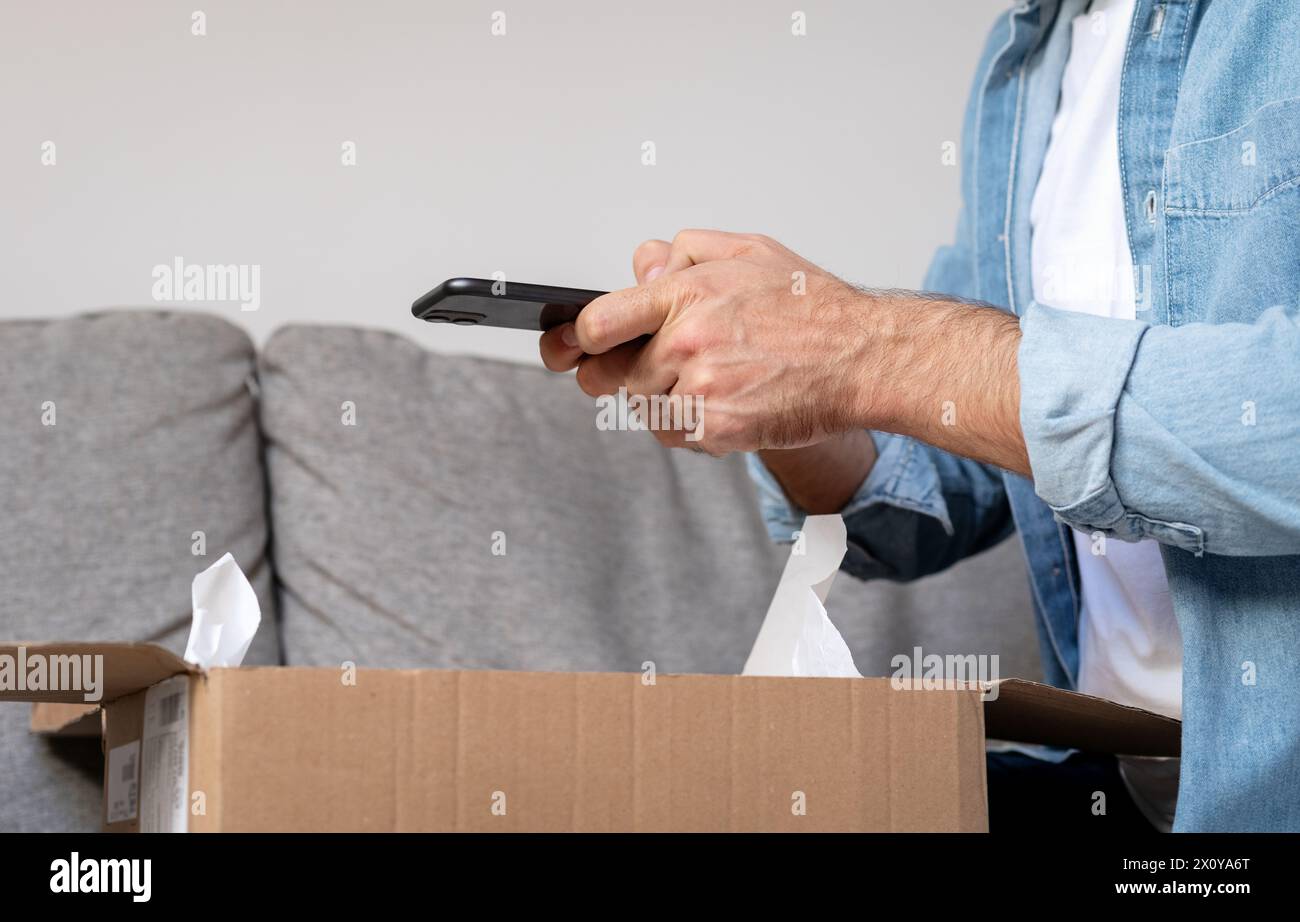 Ein Mann packt Paketkarton aus und macht ein Foto von seinem Kauf, Internetbestellung. Person, die Feedback mit seinem Smartpho an den Online-Shop weitergibt Stockfoto