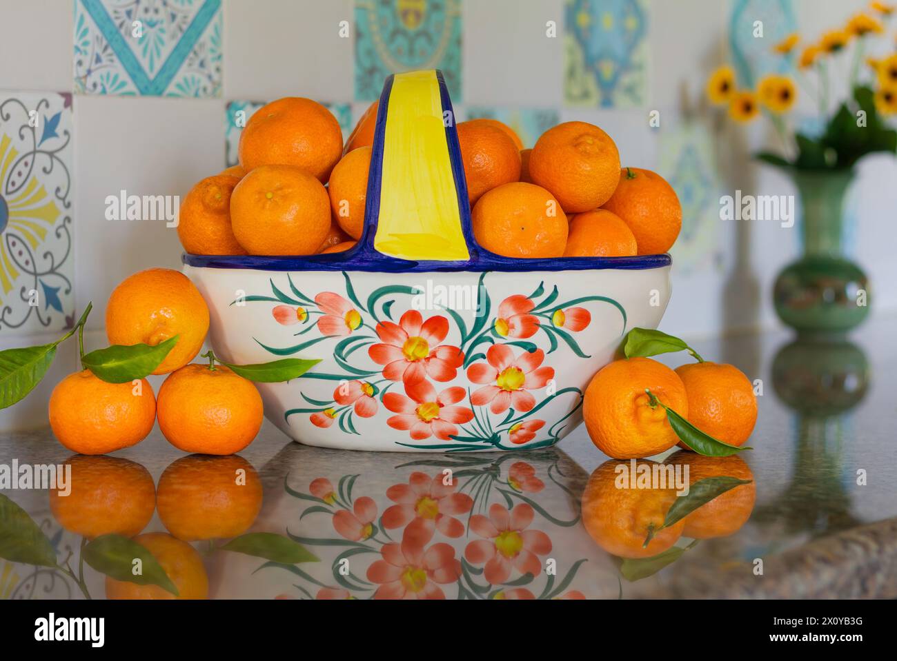 Clemantinen in einer Keramikschale auf einer reflektierenden Arbeitsplatte aus Granit mit Blumenvase Stockfoto