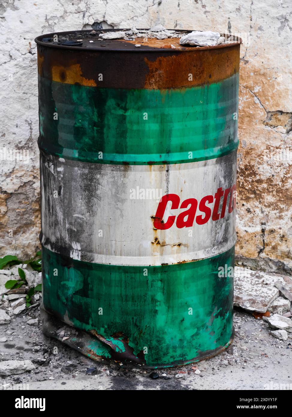 Grünes und weißes Ölfass mit rotem Castrol-Logo auf weißem Hintergrund, an einer Steinmauer stehend. Durch Feuer verfärbt und angetrübt. Stockfoto