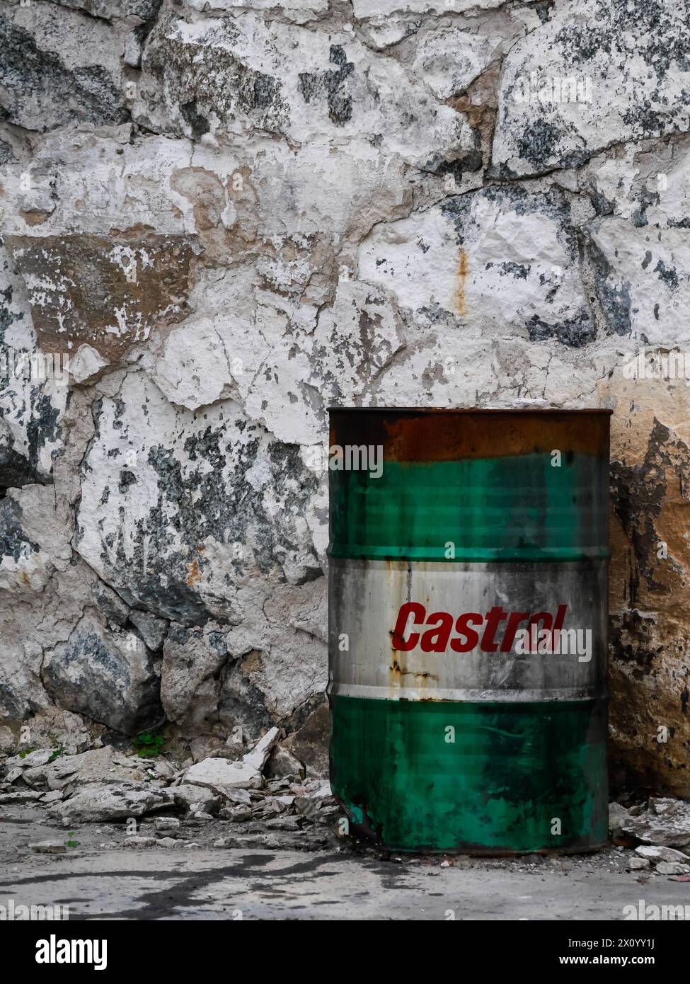 Grünes und weißes Ölfass mit rotem Castrol-Logo auf weißem Hintergrund, an einer Steinmauer stehend. Durch Feuer verfärbt und angetrübt. Stockfoto