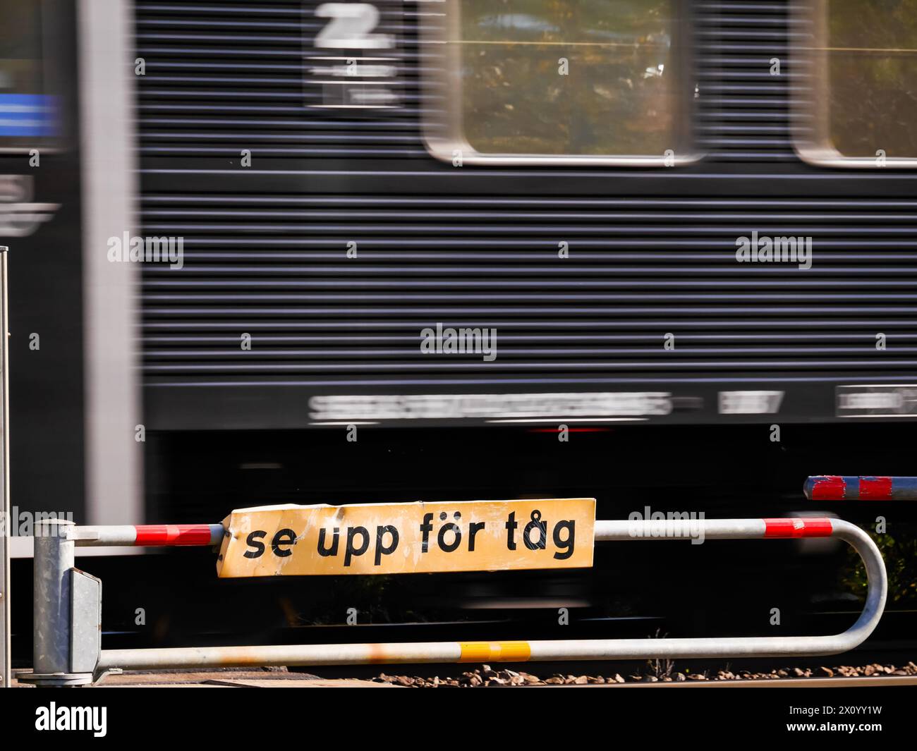 Schwarzer Zug am Bahnübergang vorbei mit einem Warnschild mit der Aufschrift „Acht auf Züge“ in schwedischer Sprache, „Varning för tåg“. Stockfoto