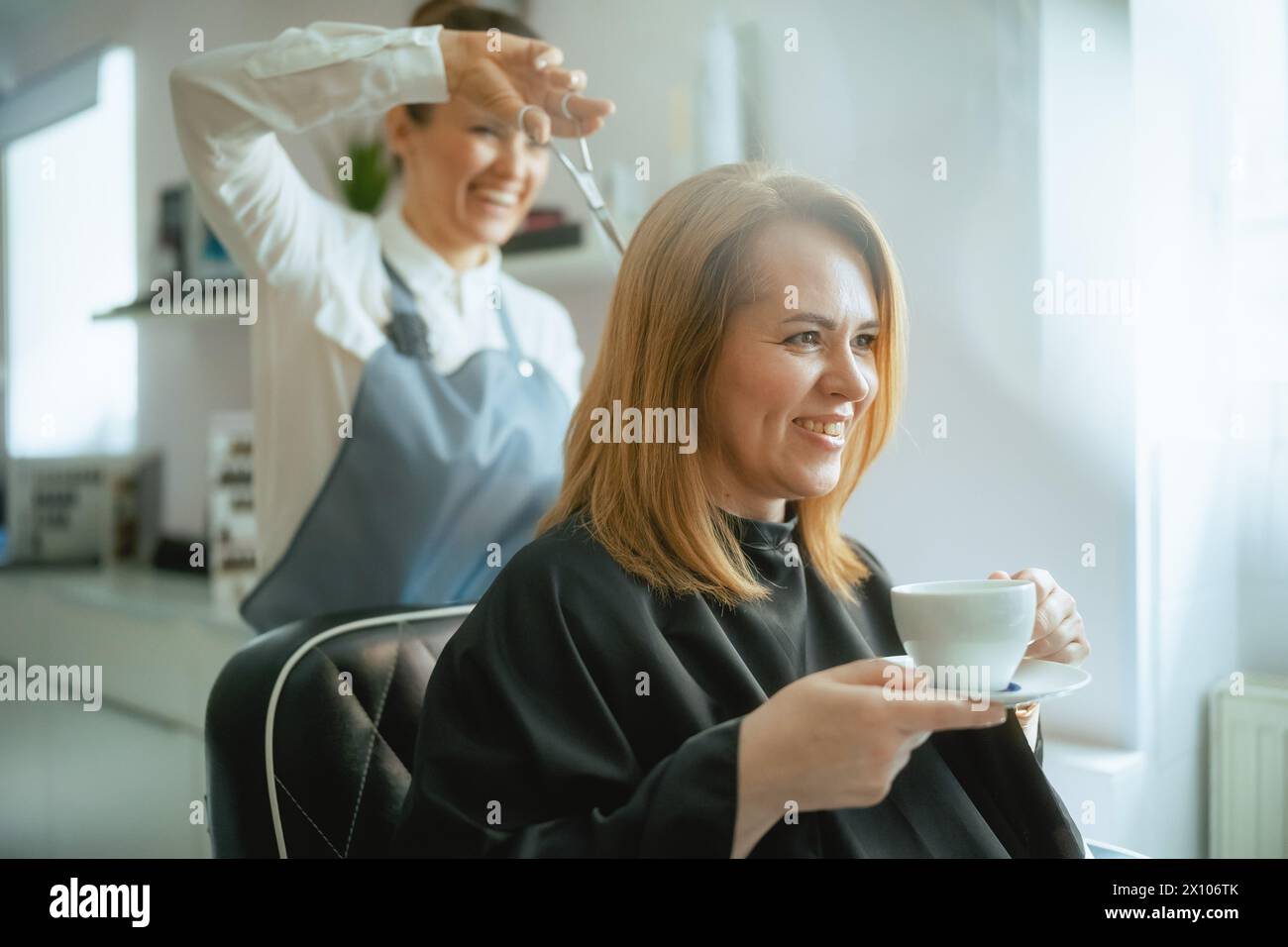 Angestellte des Friseursalons mittleren Alters in einem modernen Friseurstudio schneidet die Haare mit einer Schere und entspannt die Kundin mit einer Tasse Tee. Stockfoto