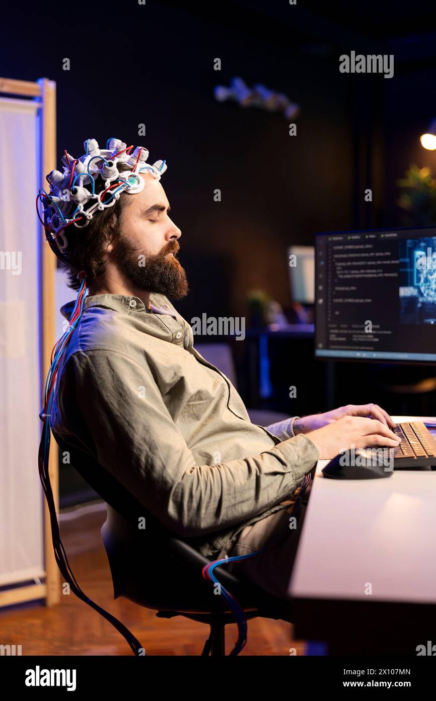 Mann mit EEG-Headset, der auf der Tastatur schreibt und Code schreibt, sodass er den Geist in die virtuelle Welt des Computers transferieren kann. Transhumanist schließt die Augen, transzendiert, wird eins mit KI Stockfoto