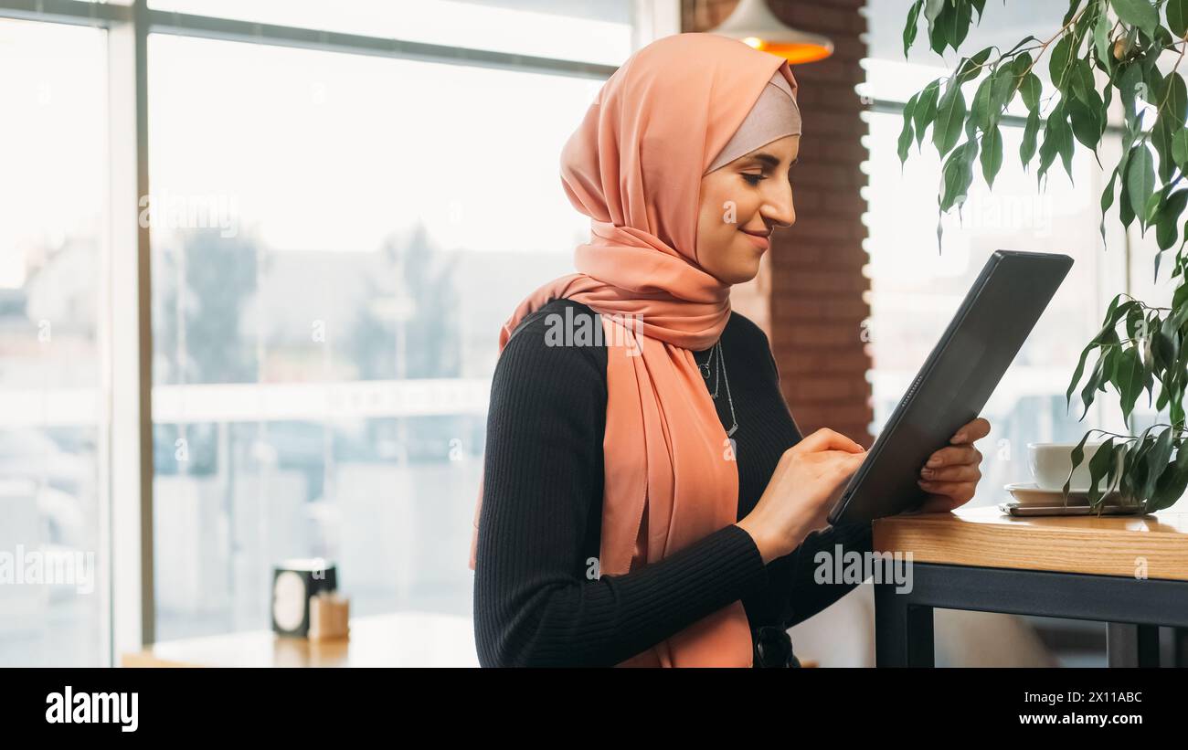 Cafe Freelance. Internetbenutzer. Fröhliche Frau im Hijab chatten auf einem digitalen Tablet im Internet surfen in sozialen Netzwerken im gemütlichen Café-Shop Kopierraum. Stockfoto