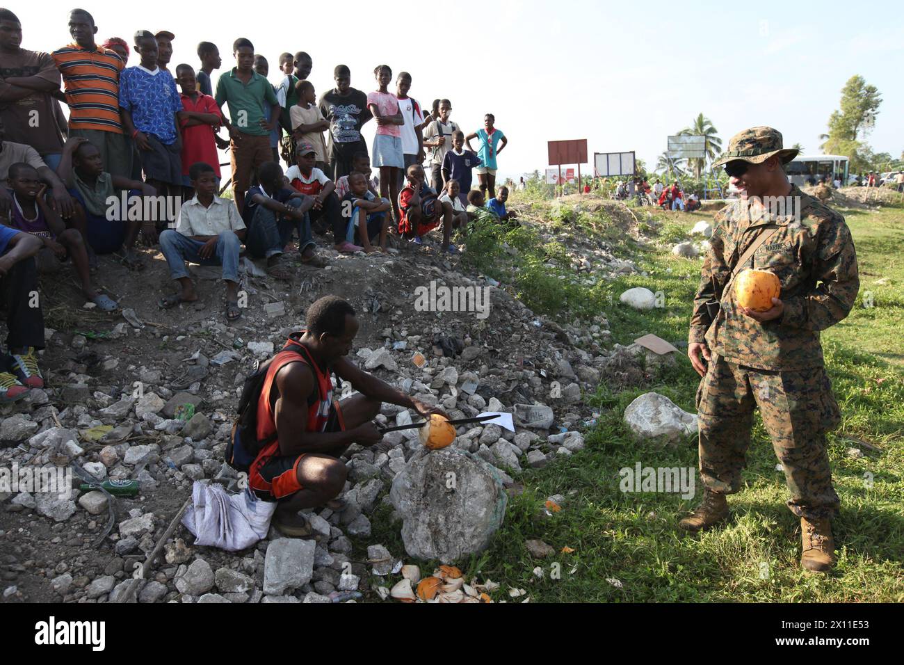 Clifford Sajous, ein Teamleiter und Linguist mit dem Battalion Landing Team, 3. Bataillon, 2. Marine Regiment, 22. Marine Expeditionary Unit, beobachtet, wie ein lokaler Haitianer während einer Wasser- und Nahrungsversorgungsmission in Leogane frische Kokosnüsse für Marines hackt 2010. Stockfoto