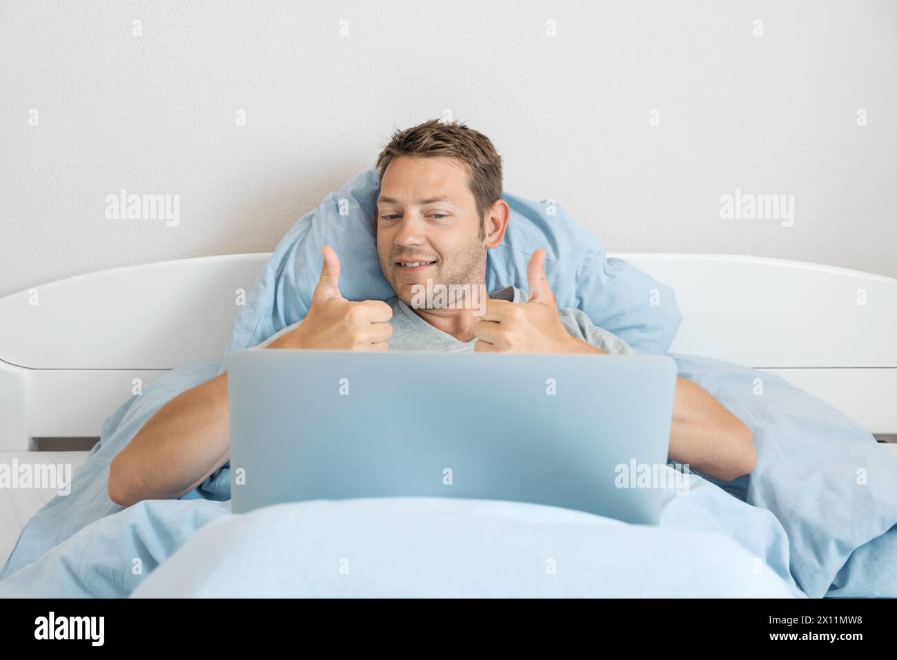 Junger glücklicher Mann kommuniziert per Videoanruf mit Freunden zeigt Daumen nach oben Geste, während er im Bett liegt und zu Hause an einem Laptop arbeitet. Fröhlich Stockfoto