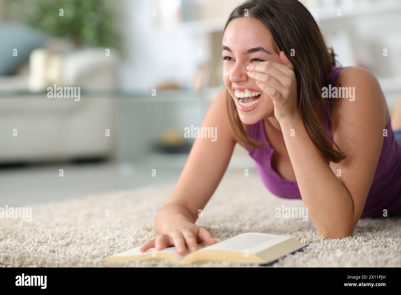 Glückliche Frau lacht urkomisch, liest ein komödiantisches Papierbuch, das zu Hause auf dem Boden liegt Stockfoto