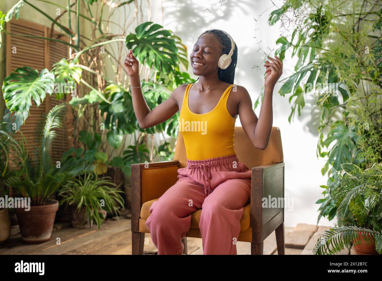 Fröhliche schwarze Frau, die auf einem Stuhl sitzt und Lieblingsmusik hört, umgeben von exotischen Zimmerpflanzen. Stockfoto