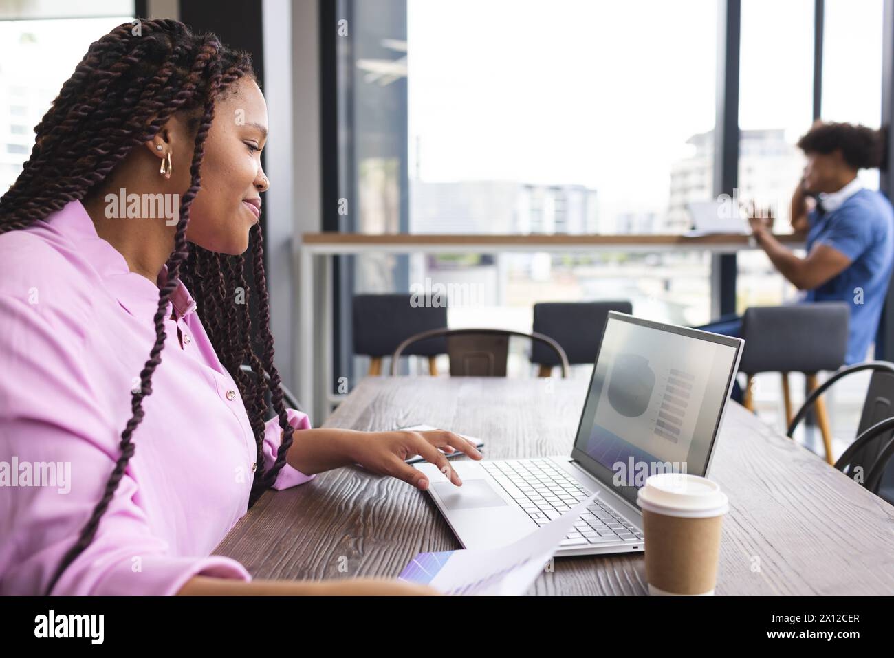 Eine junge Frau mit geflochtenen Haaren arbeitet in einem modernen Geschäftsbüro an einem Laptop Stockfoto