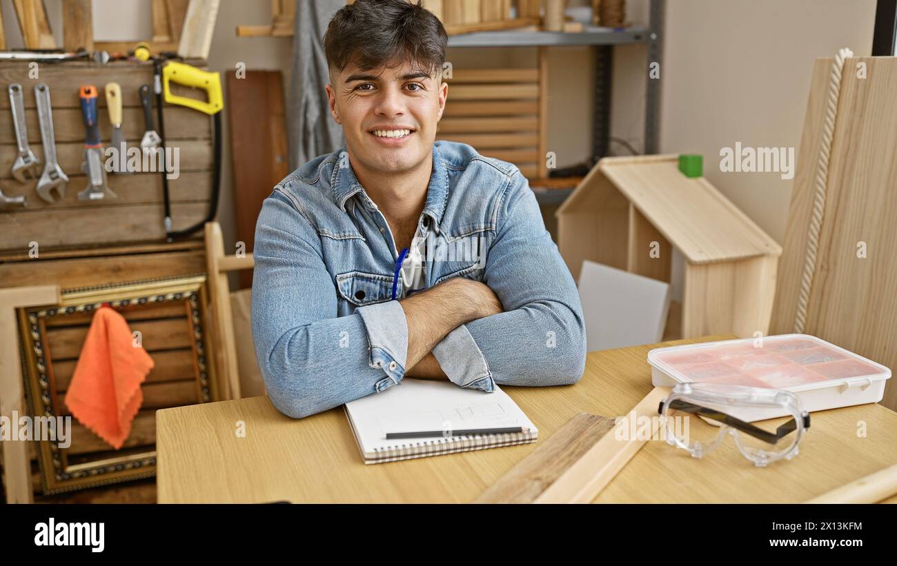 Attraktiver junger hispanischer Mann, ein professioneller Zimmermann, sitzt mit gekreuzten Armen und lächelt bei der Arbeit in seinem belebten Holzwerkstudio. Stockfoto