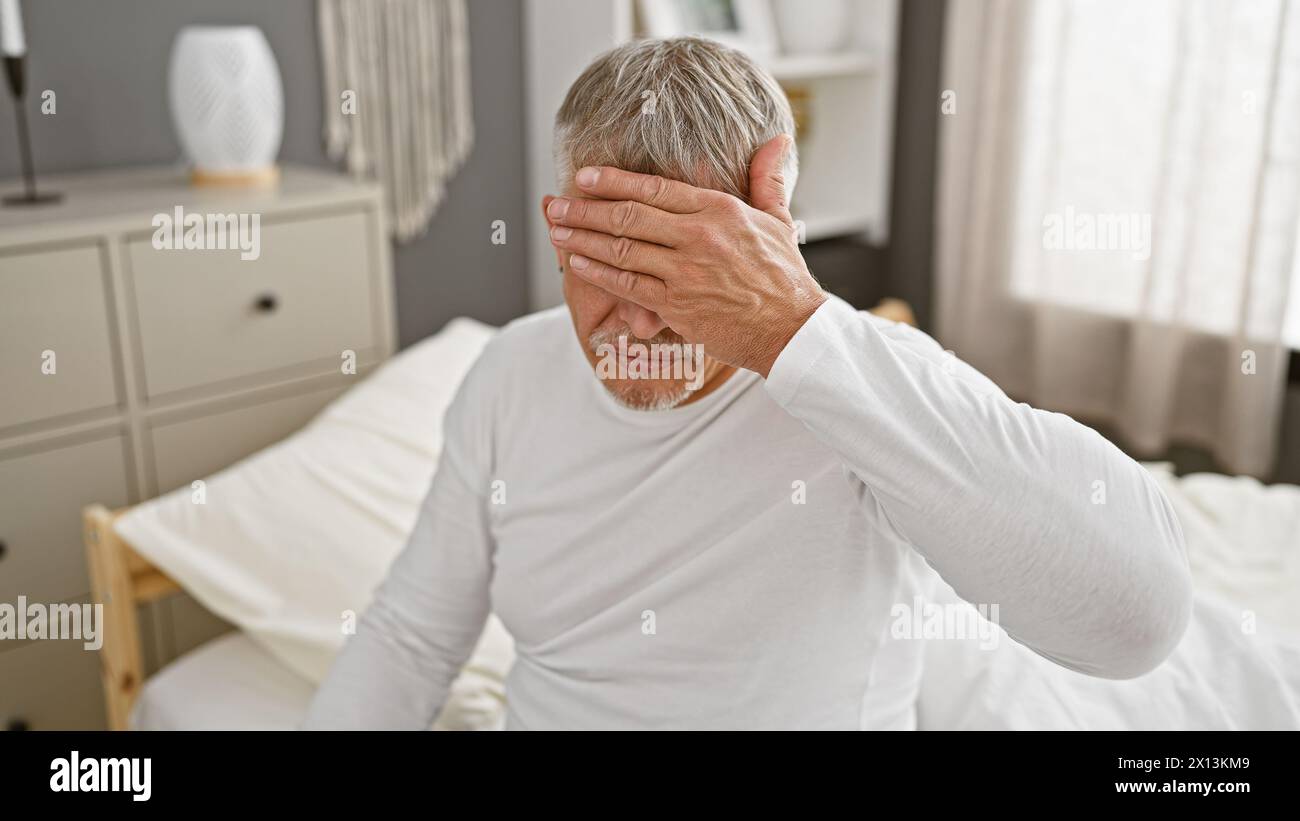 Ein beunruhigter älterer Mann mit grauen Haaren sitzt in einem Schlafzimmer und bedeckt sein Gesicht mit der Hand. Stockfoto