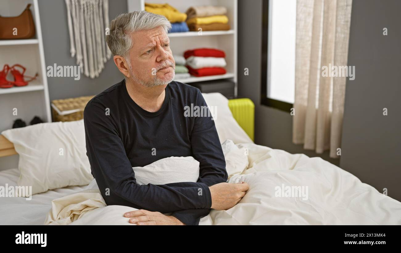 Ein besinnlicher Mann mittleren Alters mit grauen Haaren, der in einem gepflegten Bett in einem modernen Schlafzimmer sitzt und Nachsicht ausdrückt Stockfoto