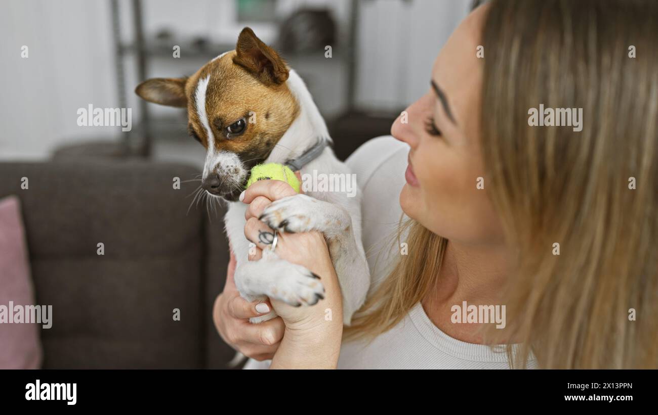 Eine junge blonde Frau spielt liebevoll mit ihrem kleinen Hund drinnen und zeigt eine Bindung zwischen Besitzer und Haustier. Stockfoto