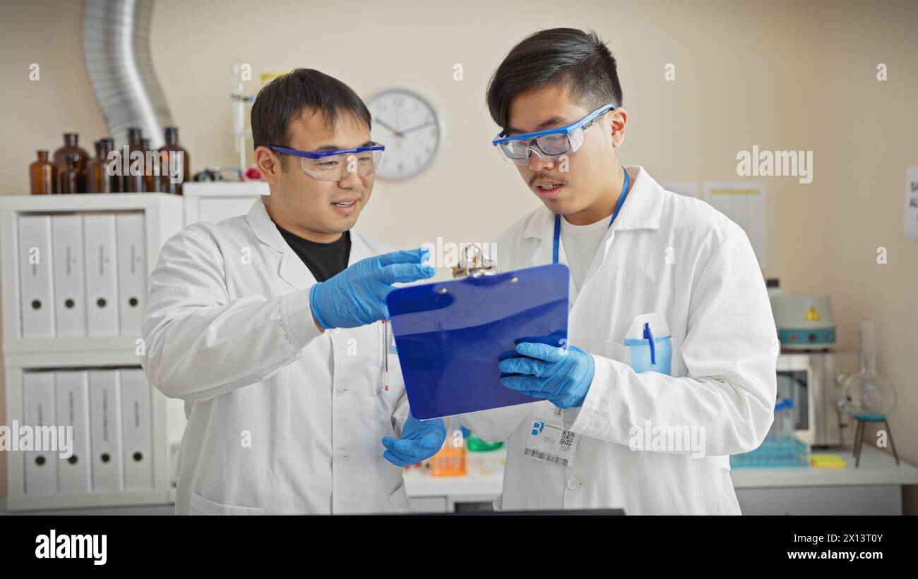 Zwei Männer in Labormänteln arbeiten in einem wissenschaftlichen Labor zusammen und diskutieren über ein Klemmbrett. Stockfoto