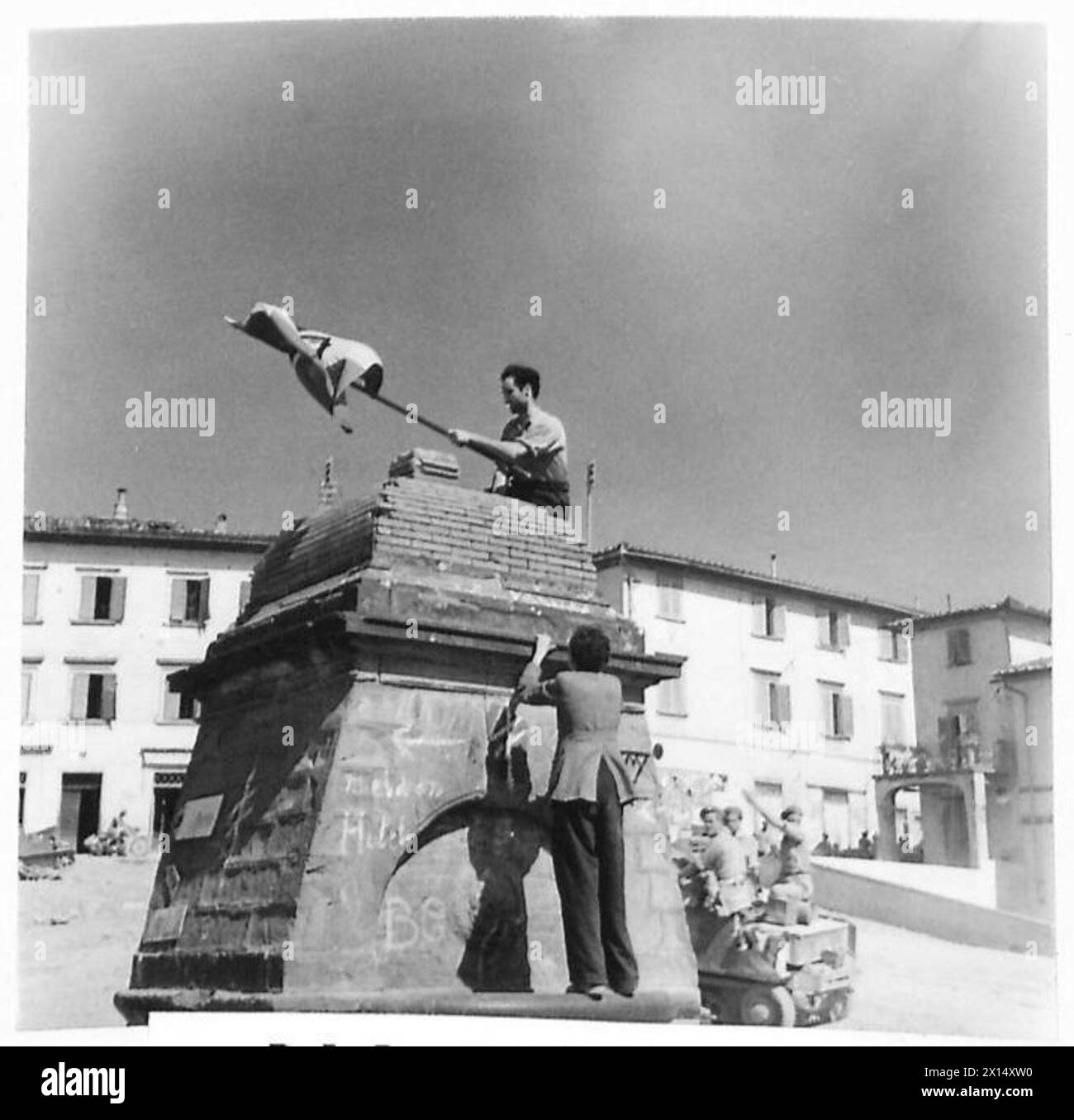 Beim Betreten einer Stadt beobachten britische Truppen der achten Armee Italiener, die eine Nationalflagge auf einem Brunnen auf dem Hauptplatz platzieren, was lokale Interaktionen hervorhebt. Stockfoto