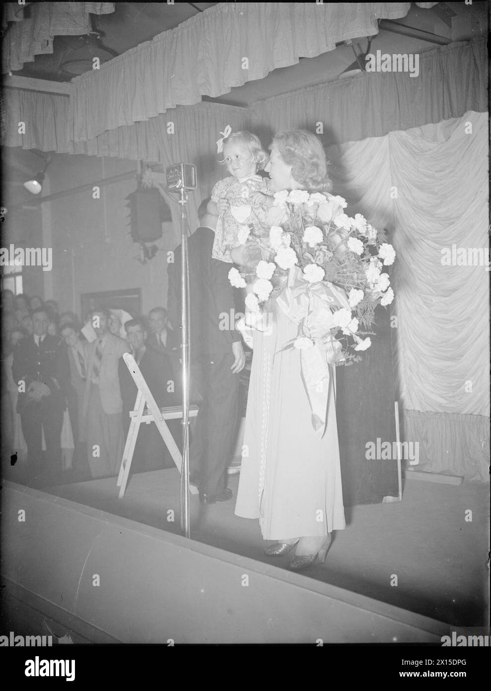 GRACIE FIELDS SINGT IN Einer MUNITIONSFABRIK, Großbritannien, 1941 - Singer Gracie Fields steht am Mikrofon auf einer Bühne einer Kampfmittelfabrik irgendwo in Großbritannien. In ihren Armen hält sie die Tochter eines Kriegsarbeiters und einen großen Blumenstrauß, den ihr das kleine Mädchen gerade nach ihrer Vorstellung auf den Fabrikfeldern, Gracie, überreicht hat Stockfoto