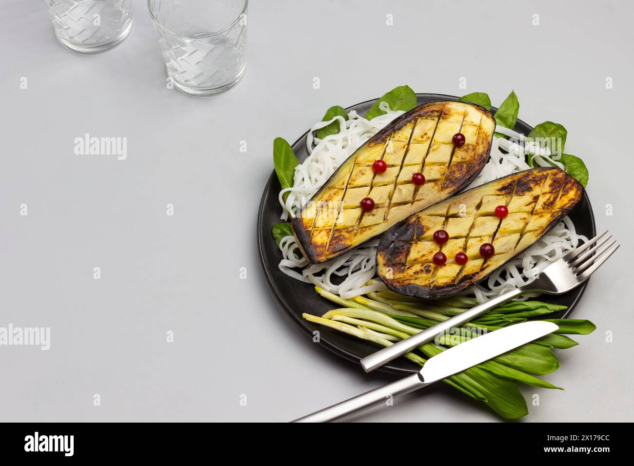 Gebackene Auberginen mit Gemüse und Reisnudeln auf schwarzer Platte. Gabel und Messer. Soße in einer Glasschale. Draufsicht Stockfoto