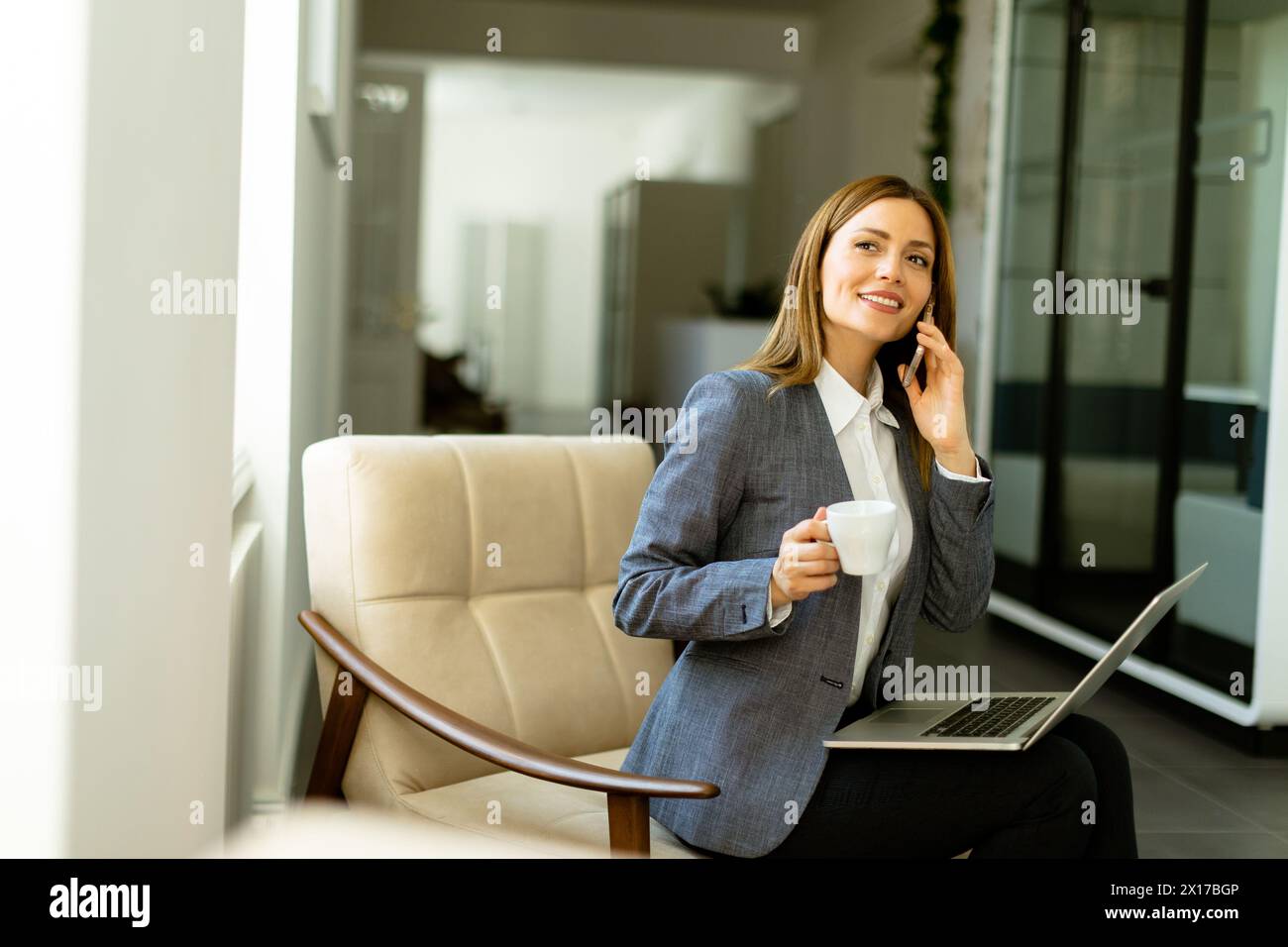 Eine Frau in Geschäftskleidung schlürft ihren Kaffee, während sie am Telefon spricht. Ihr Laptop ist bereit für die täglichen Aufgaben. Stockfoto