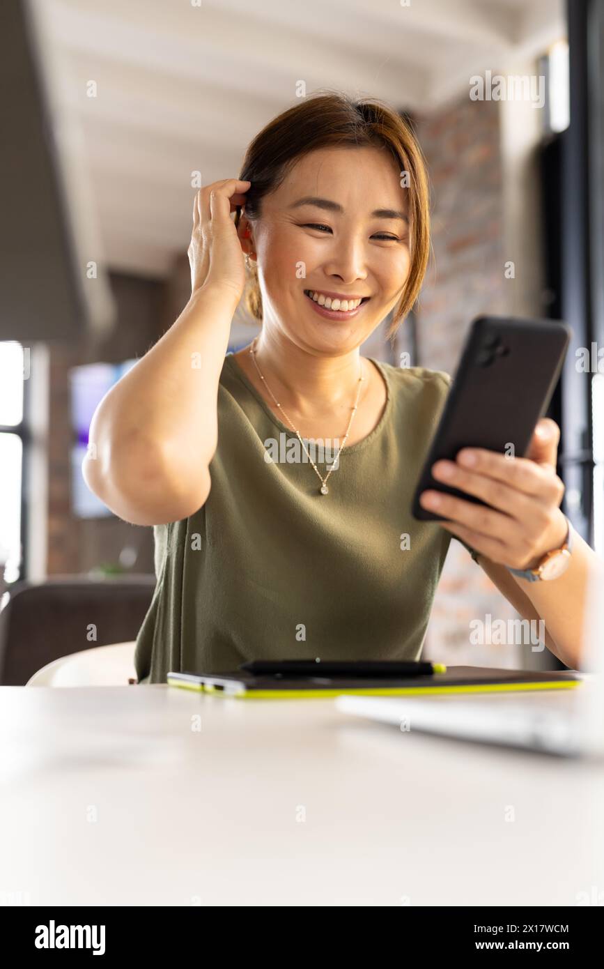 Asiatische reife professionelle Frau, die in einem modernen Geschäftsbüro lächelnd auf das Smartphone schaut Stockfoto