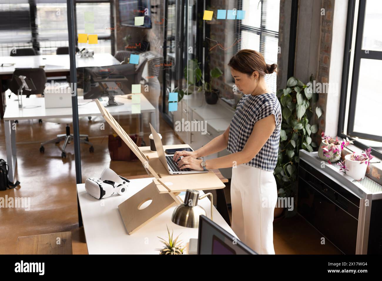 Asiatische Frau mittleren Alters, die auf einem Laptop am Stehpult in einem modernen Business-Office tippt Stockfoto