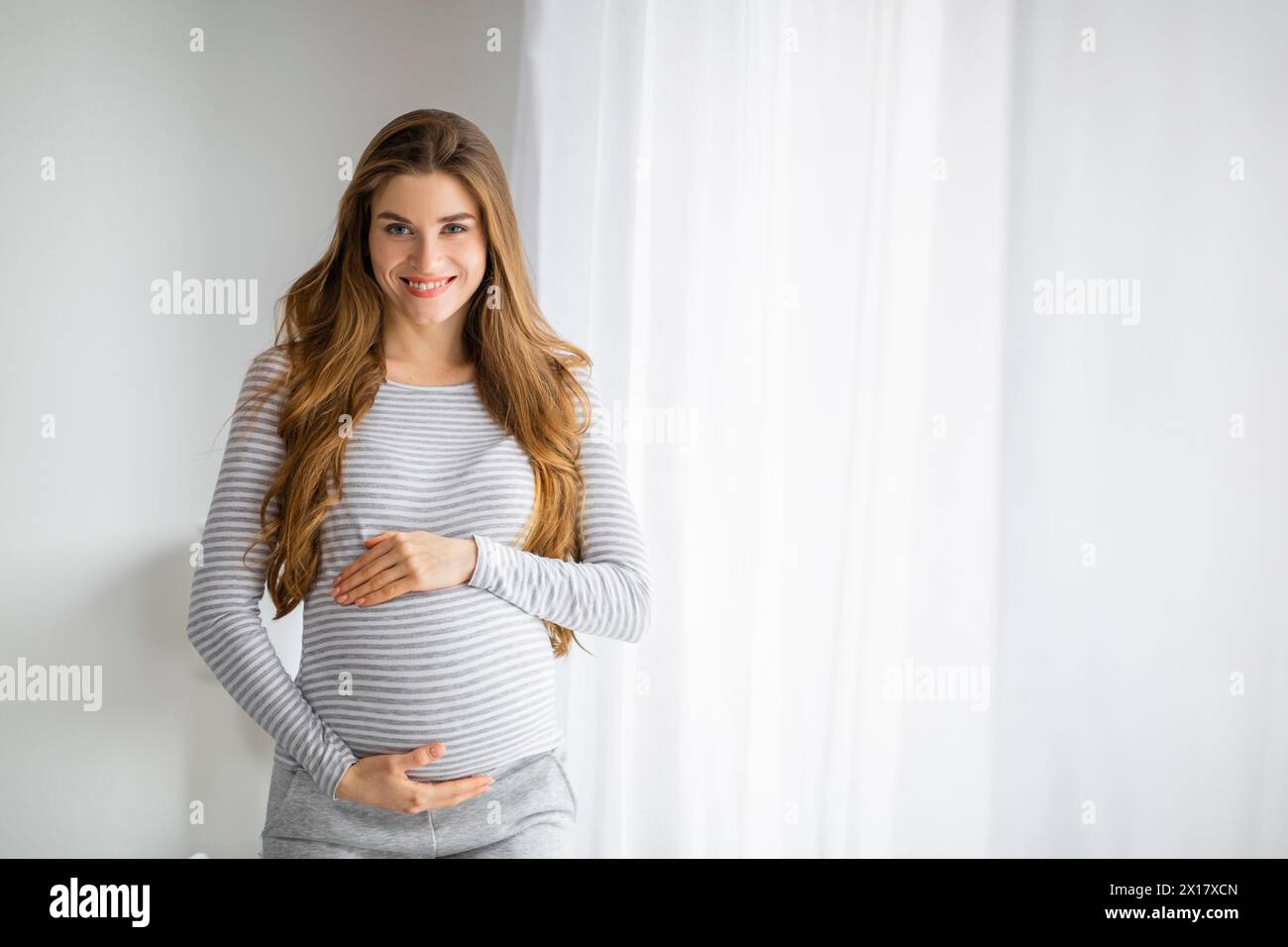 Lächelnde schwangere Frau, die am Fenster steht Stockfoto