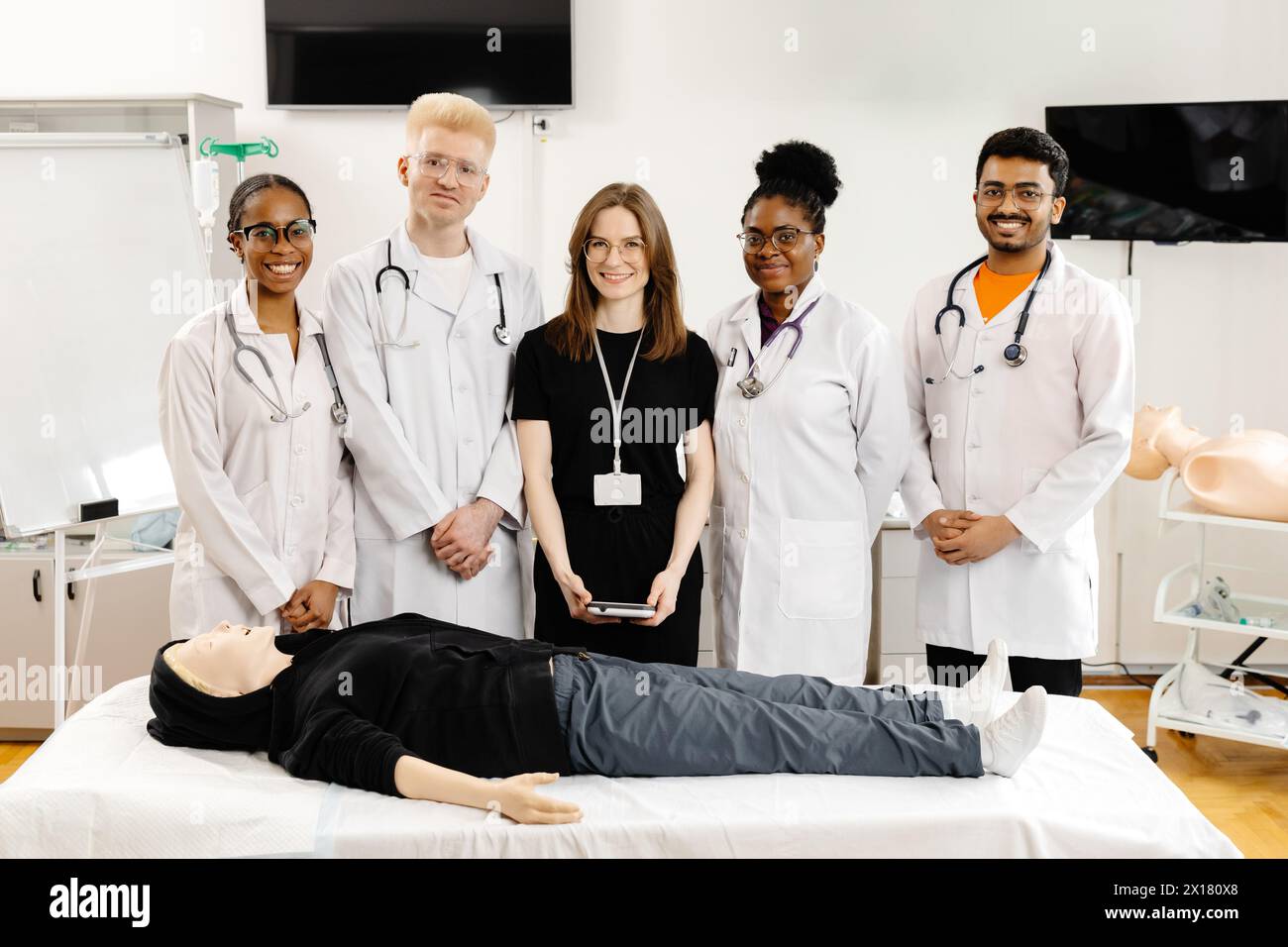 Eine Gruppe von Ärzten in weißen Mänteln, die um eine Schaufensterpuppe auf einem Krankenhausbett stehen und scheinbar über ihren Zustand oder ihre Behandlung sprechen Stockfoto