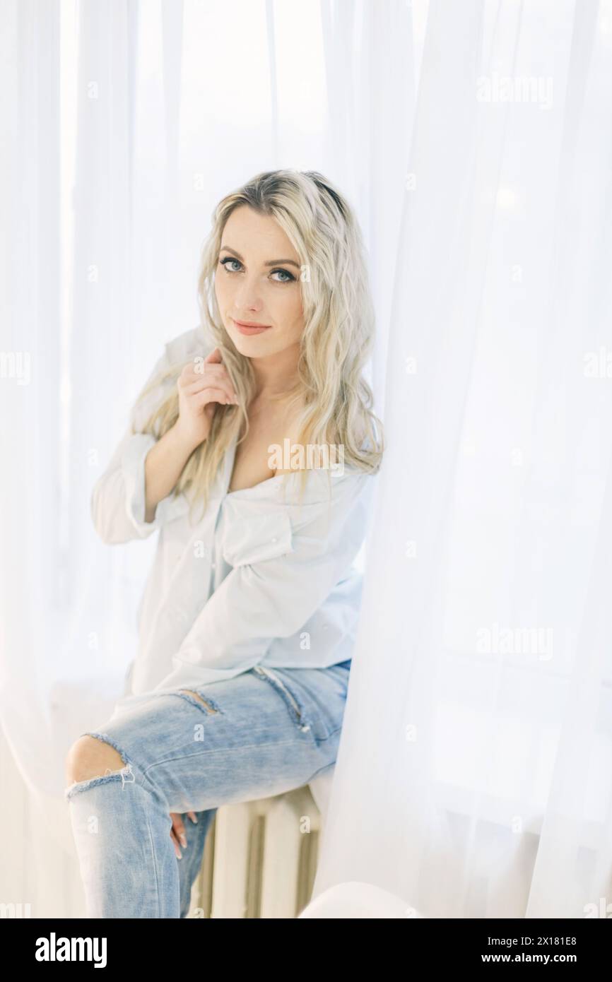 Eine Frau sitzt auf einem Heizkörper neben einem Fenster und blickt mit kontemplativem Ausdruck heraus. Stockfoto