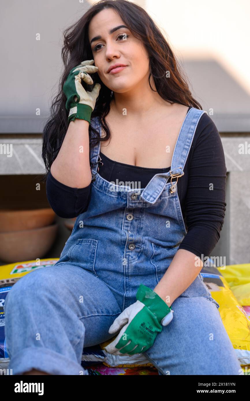 Eine Frau aus Denim-Overall und -Handschuhen sitzt lässig im Freien und hat eine entspannte Haltung Stockfoto