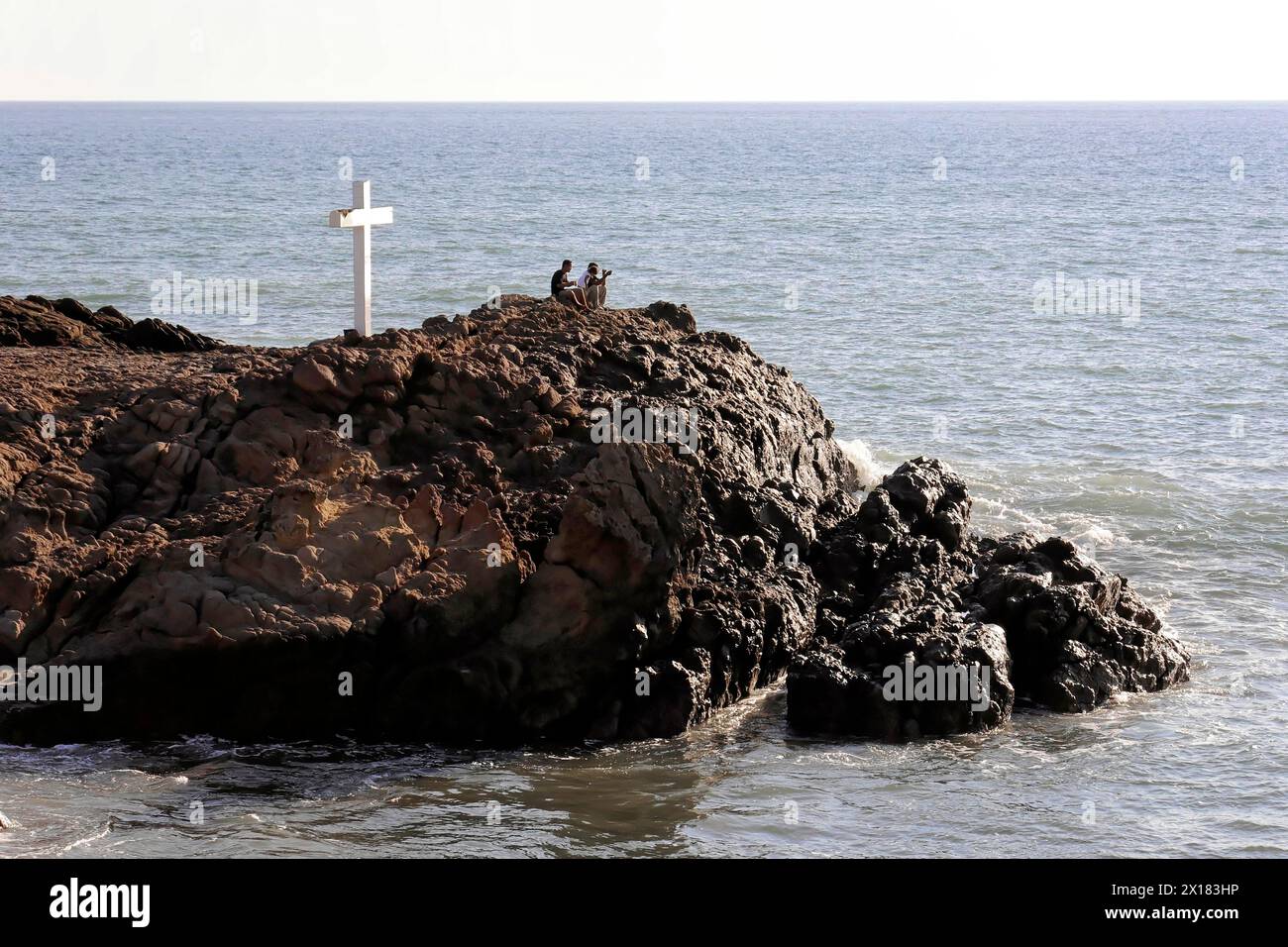 Strand in der Nähe von Poneloya, Las Penitas, Leon, Nicaragua, Mittelamerika, ein weißes Kreuz auf einem felsigen Vorsprung mit Blick auf das Meer mit zwei Personen nebenan Stockfoto