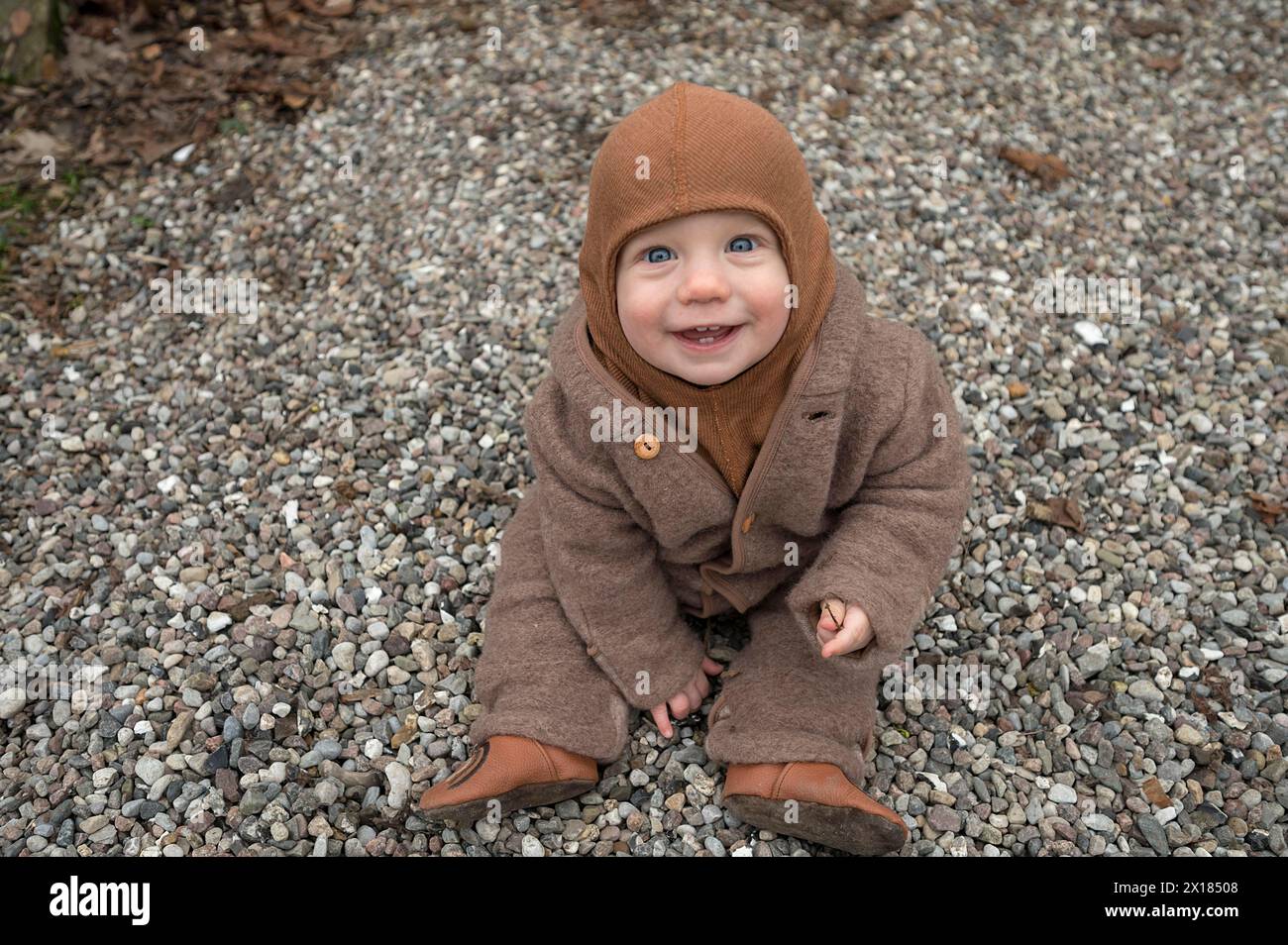Lachendes Kind, 8 Monate, warm eingewickelt, auf dem Kies sitzend und mit Steinen spielend, Mecklenburg-Vorpommern, Deutschland Stockfoto