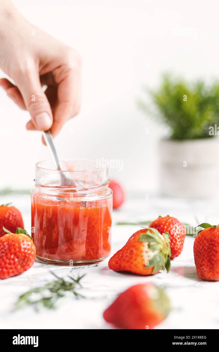 Mit einer Hand ein Glas frisch zubereitete Erdbeermarmelade umrühren. Auf dem Tisch liegen mehrere Erdbeeren Stockfoto