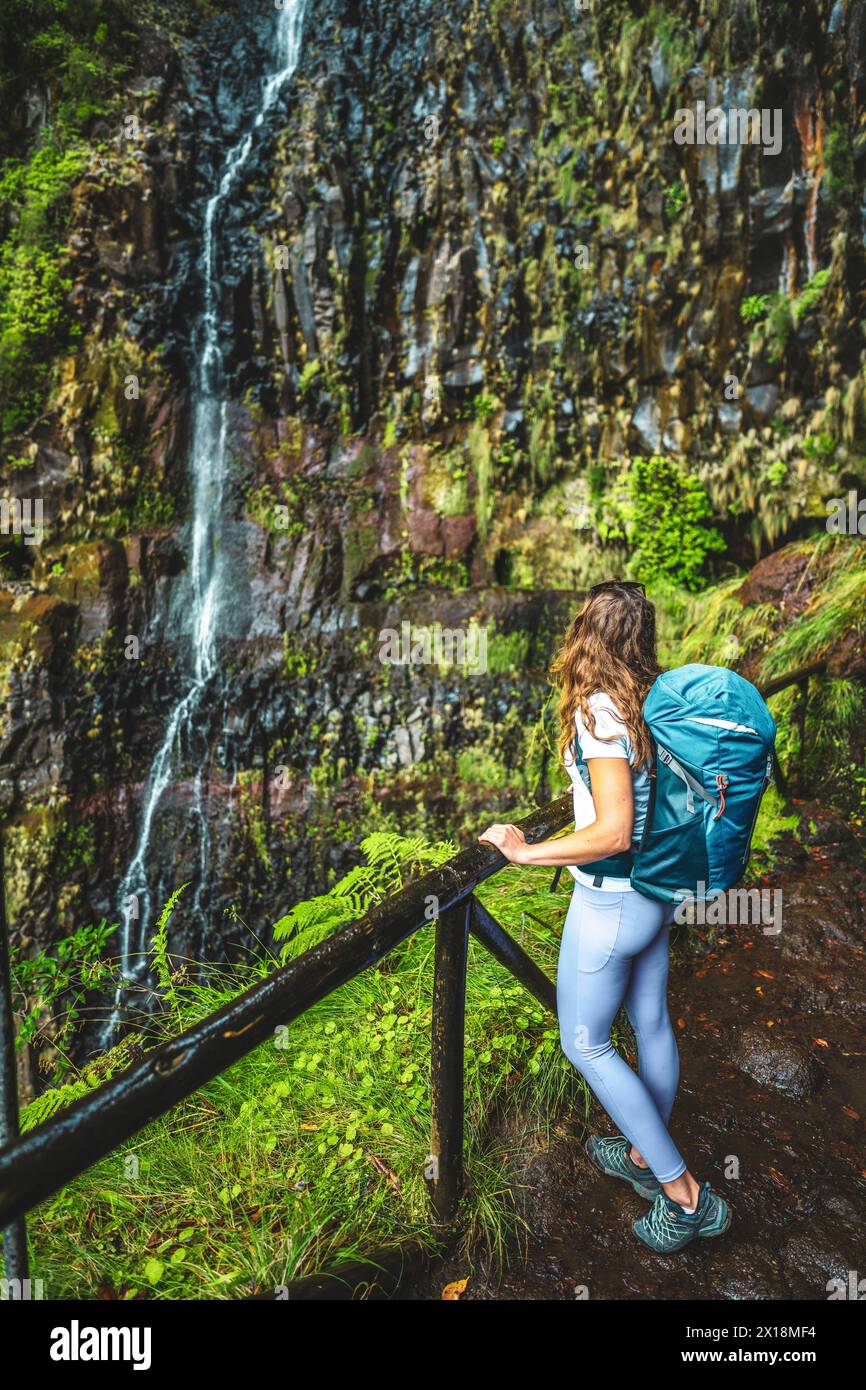 Beschreibung: Tourist mit Rucksack lehnt sich an den hölzernen Zaun des Aussichtspunkts und beobachtet den grün bewachsenen Risco-Wasserfall. 25 Fontes Wasserfälle, Madeira Stockfoto