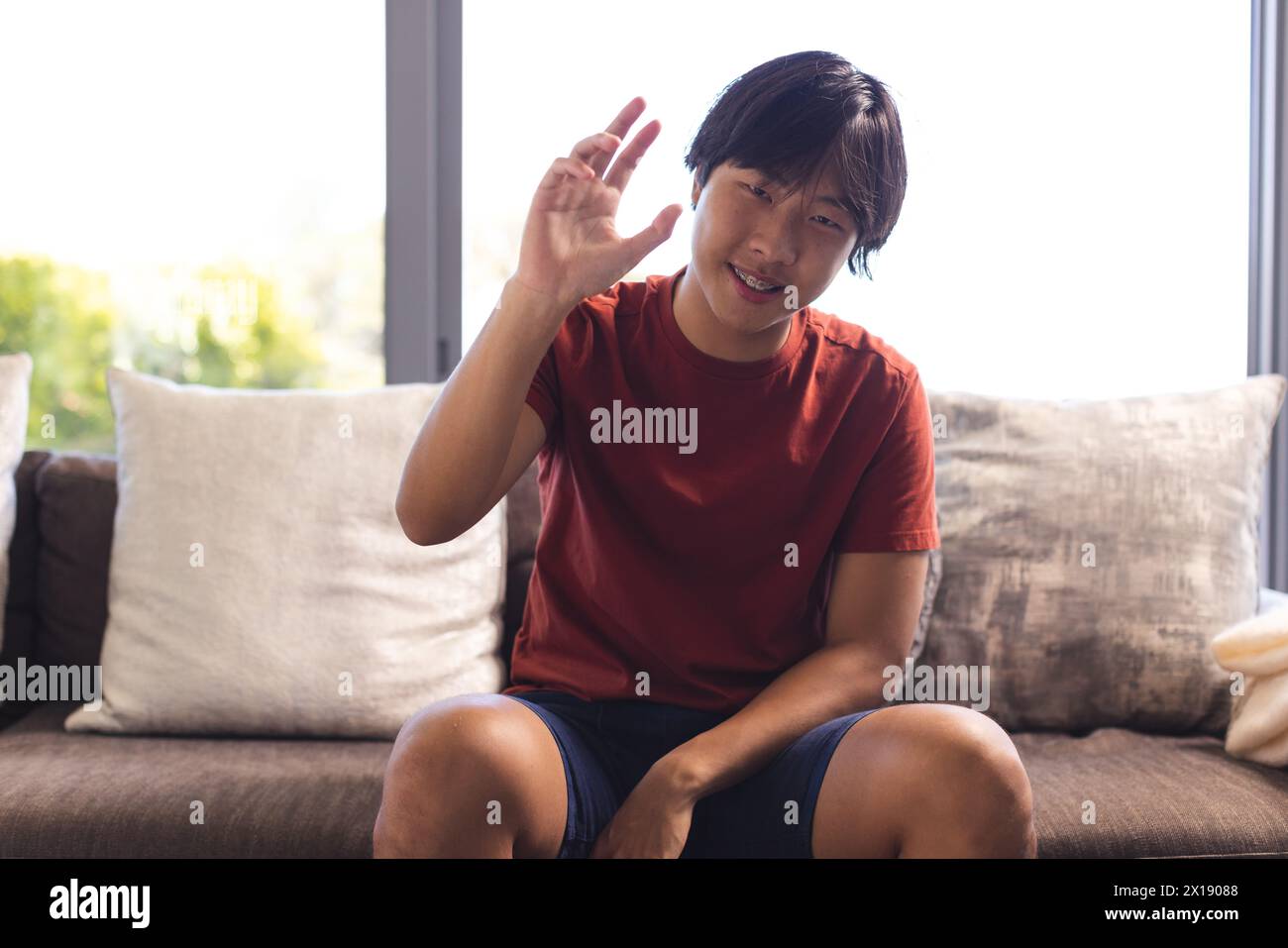 Asiatischer Teenager, der zu Hause auf der Couch sitzt und während eines Videoanrufs Handgesten macht Stockfoto