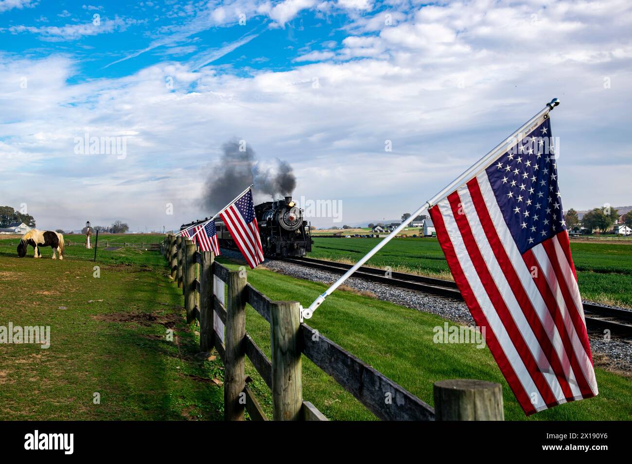 Eine alte Dampfeisenbahn zieht entlang der Landschaft, flankiert von amerikanischen Flaggen auf einem Holzzaun, die ein Gefühl nostalgischer Americana und historischer Reise weckt. Stockfoto