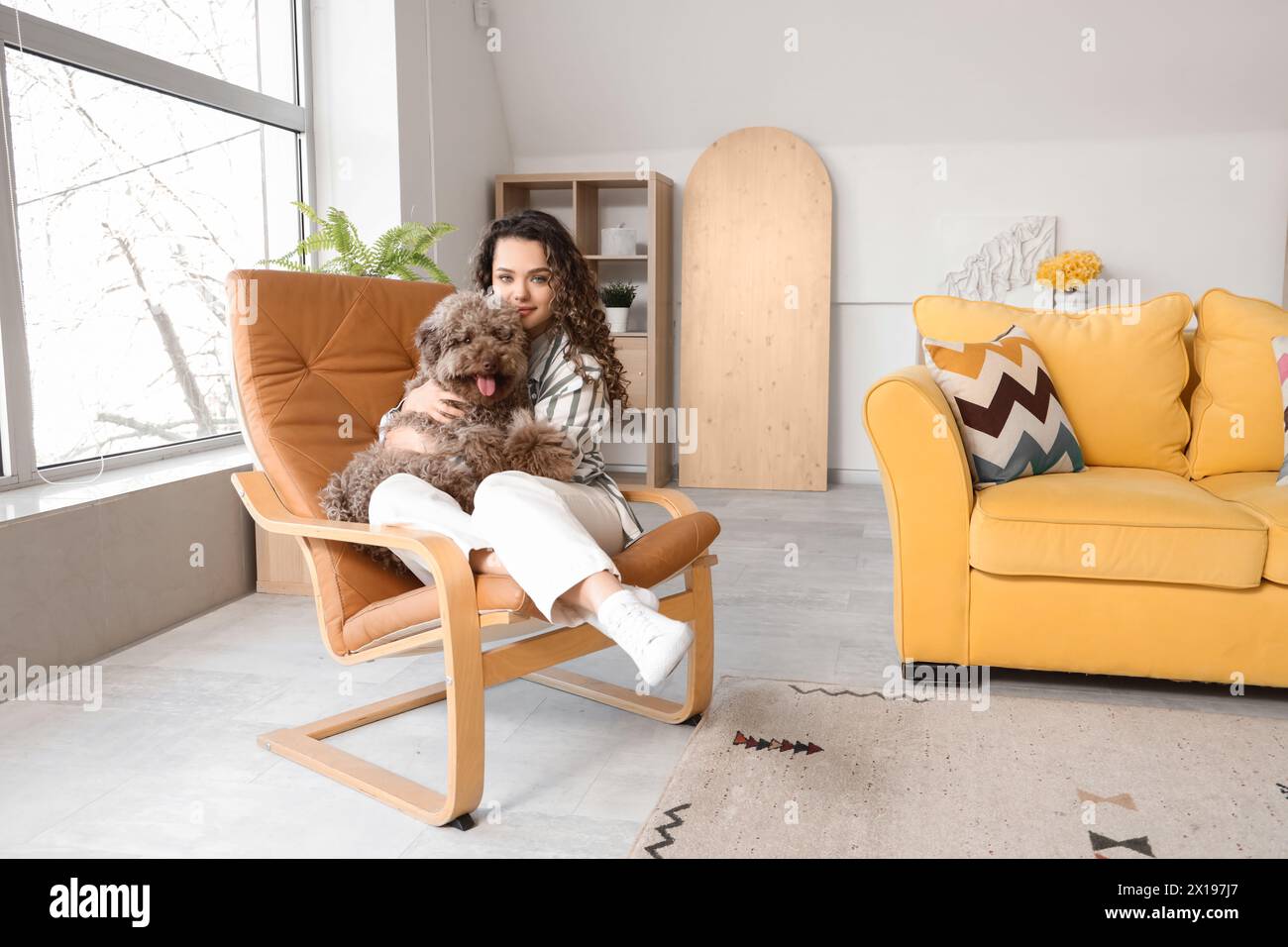 Junge Frau mit süßem Pudel, die zu Hause im Sessel sitzt Stockfoto