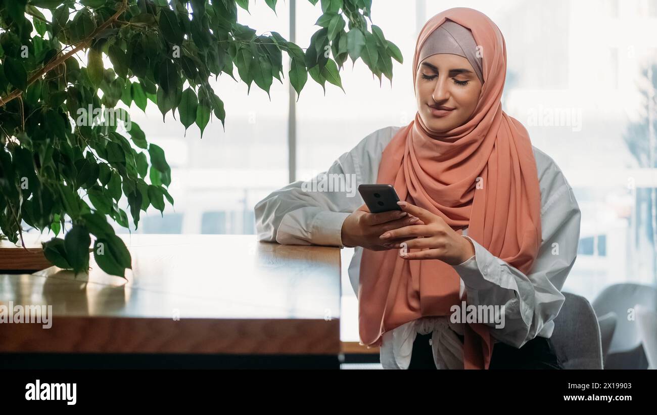 Mobiles Blogging. Freizeit im Café. Lächelnde Frau im Hijab, die soziale Medien-Nachricht auf Telefon Internet-Chat-entspannend in einem gemütlichen Café schickt. Stockfoto