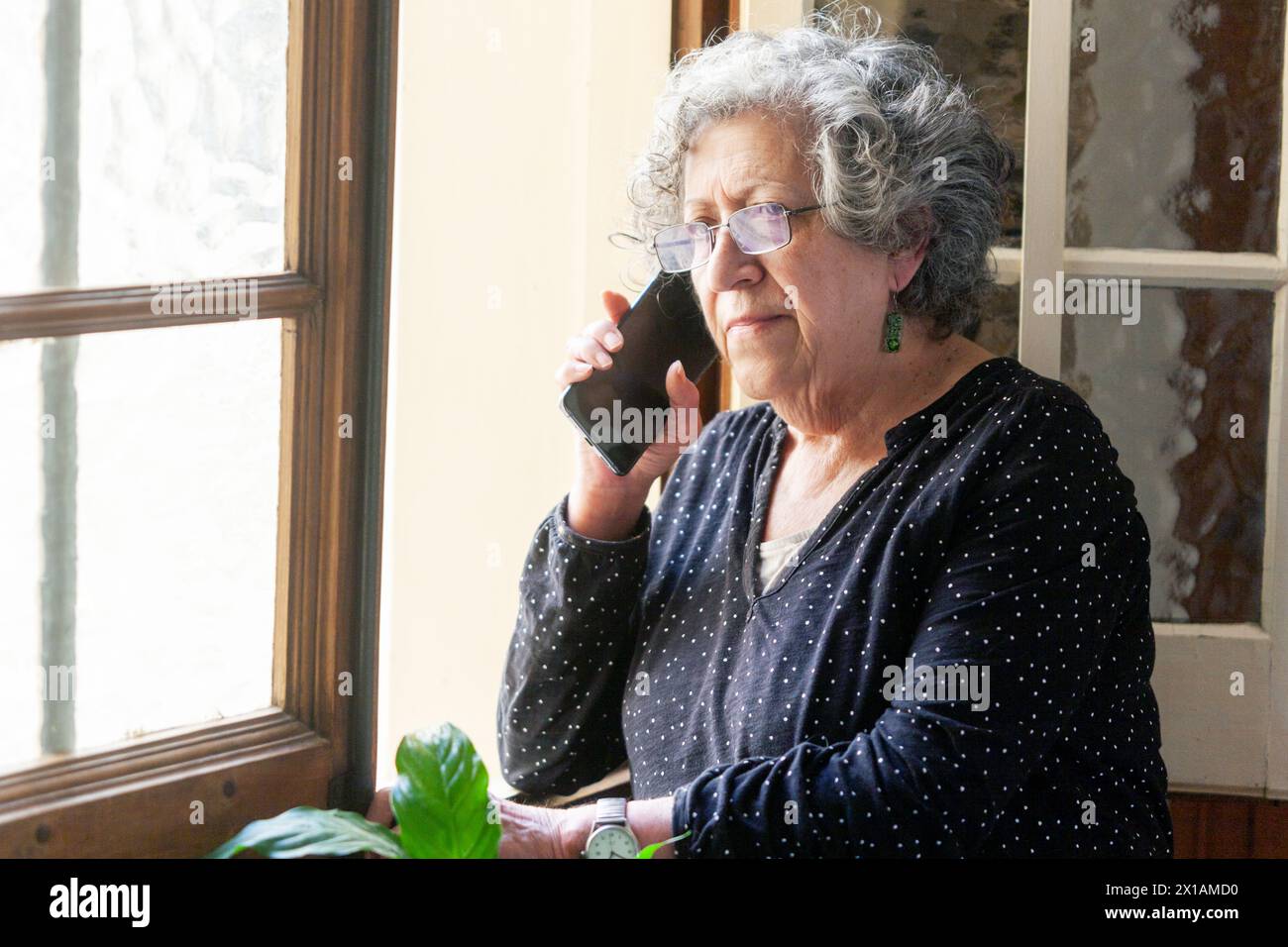 Die siebzigjährige Frau telefoniert mit ihrem Handy neben einem Fenster in ihrem Haus. Die Szene wird durch das eintretende Morgensonnenlicht beleuchtet Stockfoto