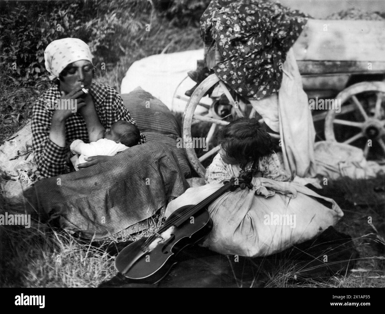 Zigeunerin, stinkende Mutter mit Säuberung an der Büste, auf Grasboden neben dem Wagenrad sitzend, davor eines weiteren Details Kind hinter dem Bündel - dabei Violine Lean - sitzend, 1930 - 19300101 PD21383 - Rechteinfo: Rights Managed (RM) Stockfoto