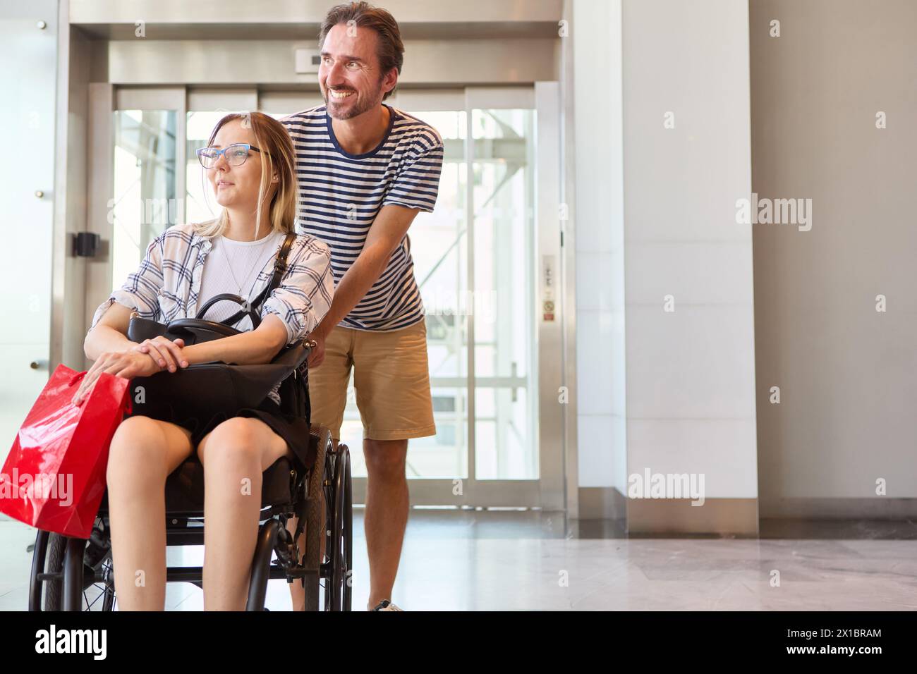 Ein lächelnder junger Mann hilft einer Frau mit einem Rollstuhl und zeigt bei einem Einkaufsausflug in ein Einkaufszentrum die Gesellschaft und Einbeziehung. Stockfoto