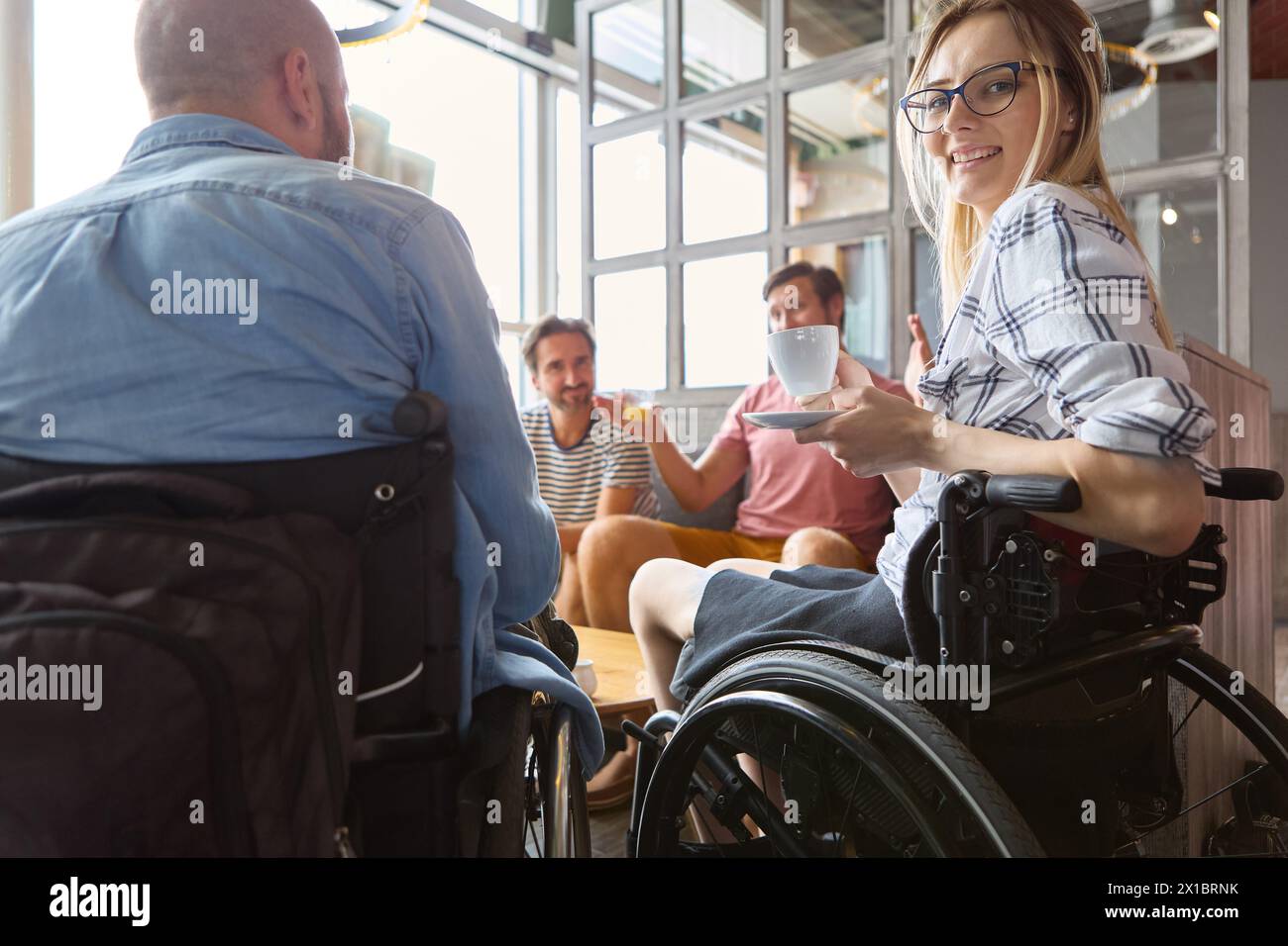 Eine Person, die einen Rollstuhl benutzt, teilt einen fröhlichen Moment mit Freunden bei einem Kaffee in einem hellen, modernen Café-Ambiente, was beispielhaft für soziale Eingliederung ist. Stockfoto