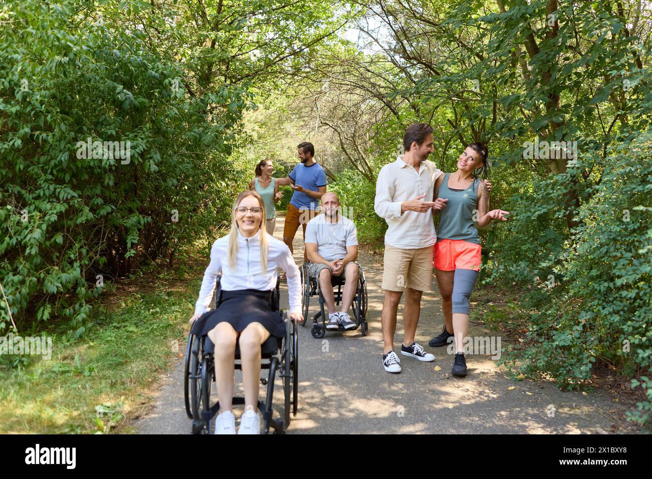 Eine vielfältige Gruppe von Freunden, darunter Rollstuhlfahrer, teilen Lachen und Gespräche während eines gemütlichen Spaziergangs in einem üppigen Park. Stockfoto