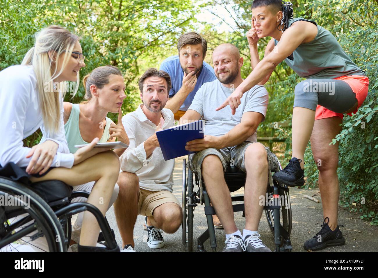 Eine Gruppe von Freunden, darunter eine Person, die einen Rollstuhl benutzt, führt eine lebhafte Diskussion in einem Park. Sie tauschen Ideen aus, verwenden digitale Tablets, Stockfoto