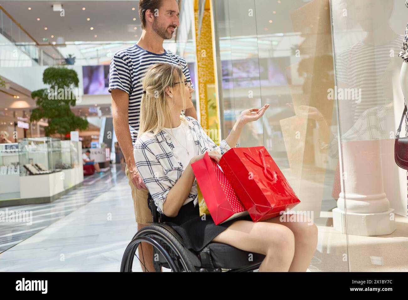 Eine Frau, die einen Rollstuhl benutzt, genießt einen Einkaufstag in einem Einkaufszentrum mit einem männlichen Begleiter, beide bewundern eine Ladenpräsentation. Stockfoto