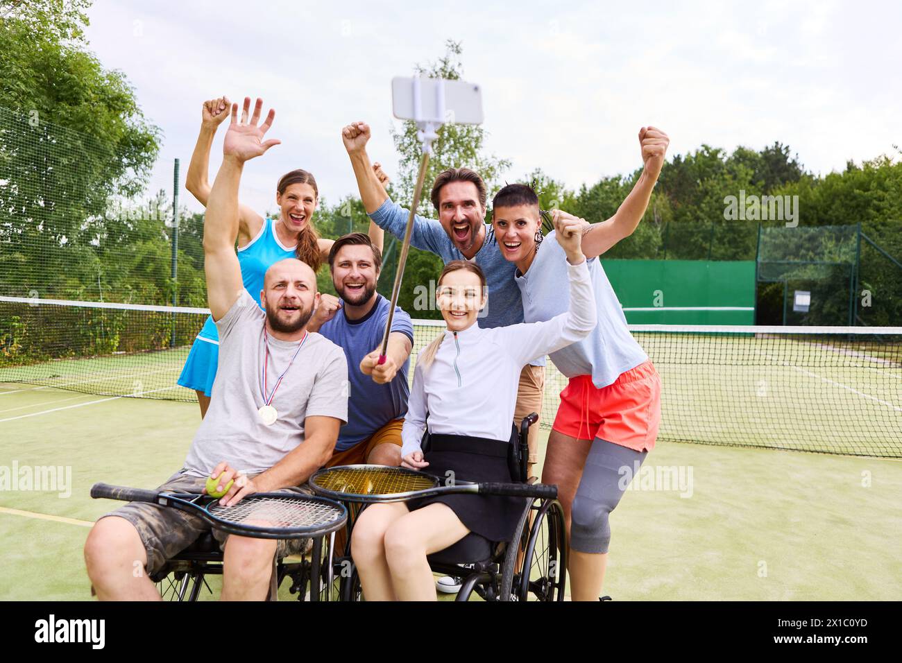 Eine fröhliche, vielfältige Gruppe von Freunden, einschließlich einer Person, die einen Rollstuhl benutzt, feiert einen Sieg auf dem Tennisplatz und zeigt Inklusion und Freundlichkeit Stockfoto