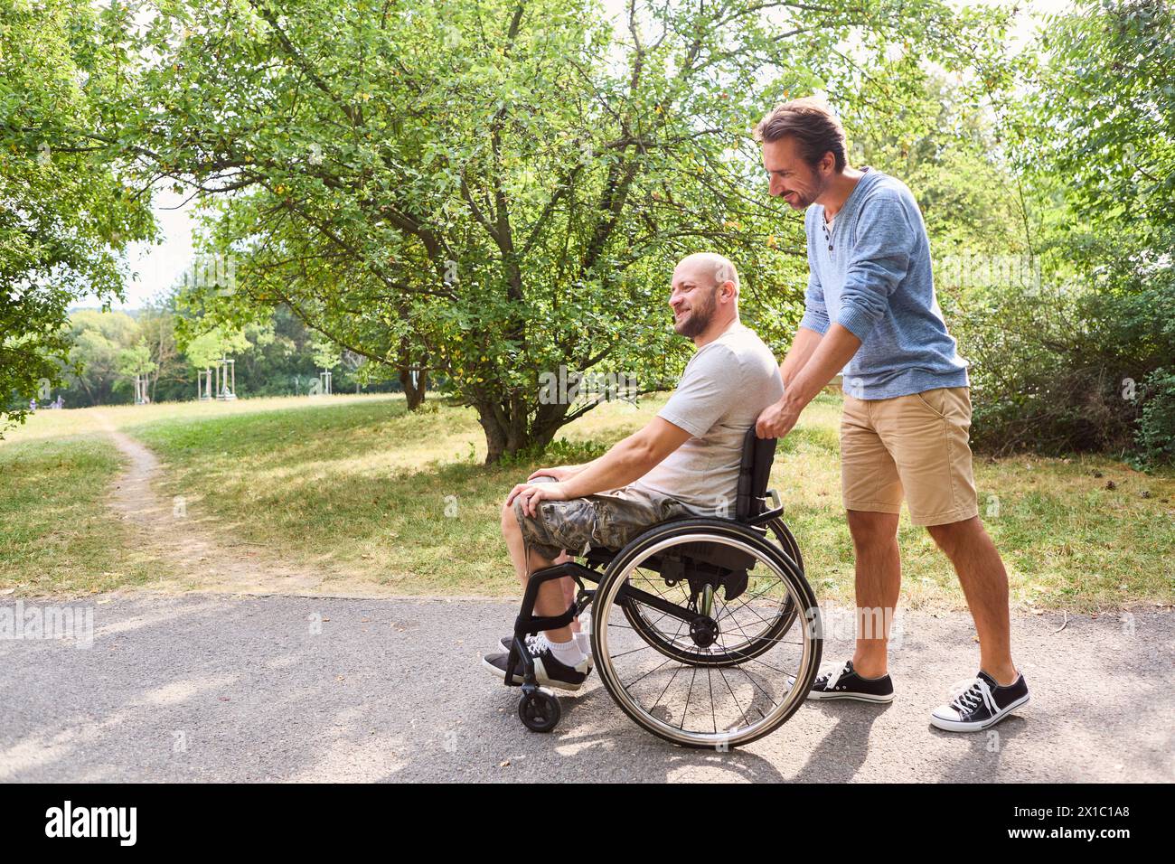 Ein Mann, der einen Rollstuhl benutzt und draußen einen freudigen Moment mit seinem Freund teilt. Sie befinden sich in einem grünen Park und betonen Freundschaft und Inklusion. Stockfoto