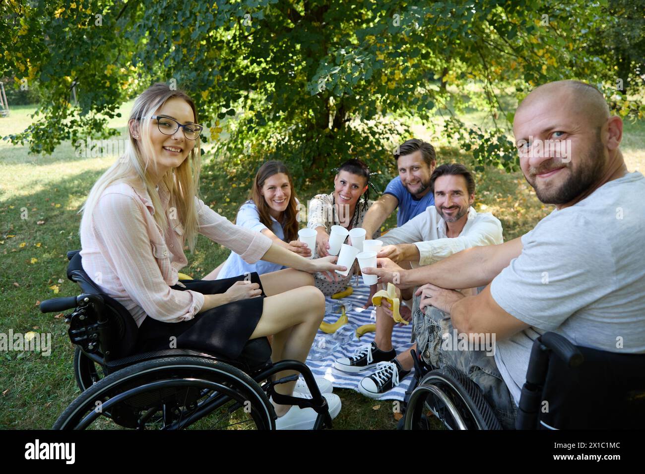 Gruppe glücklicher Freunde, die mit einer Person interagieren, die einen Rollstuhl während eines Picknicks im Park benutzt. Stockfoto