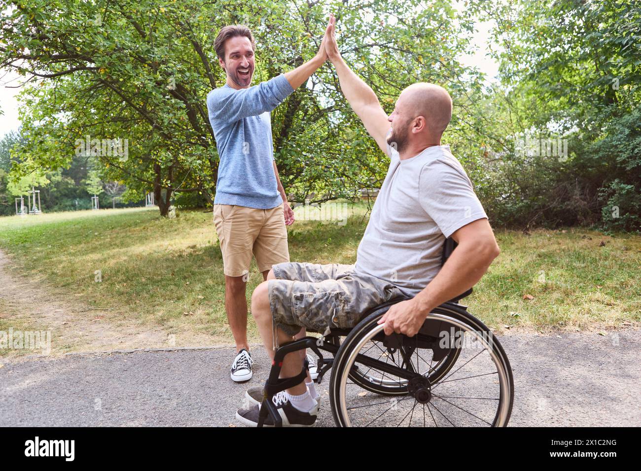 Person, die einen Rollstuhl benutzt, und sein stehender Freund High Five freudig in einem üppigen Park und zeigt Inklusion und Freundschaft. Stockfoto