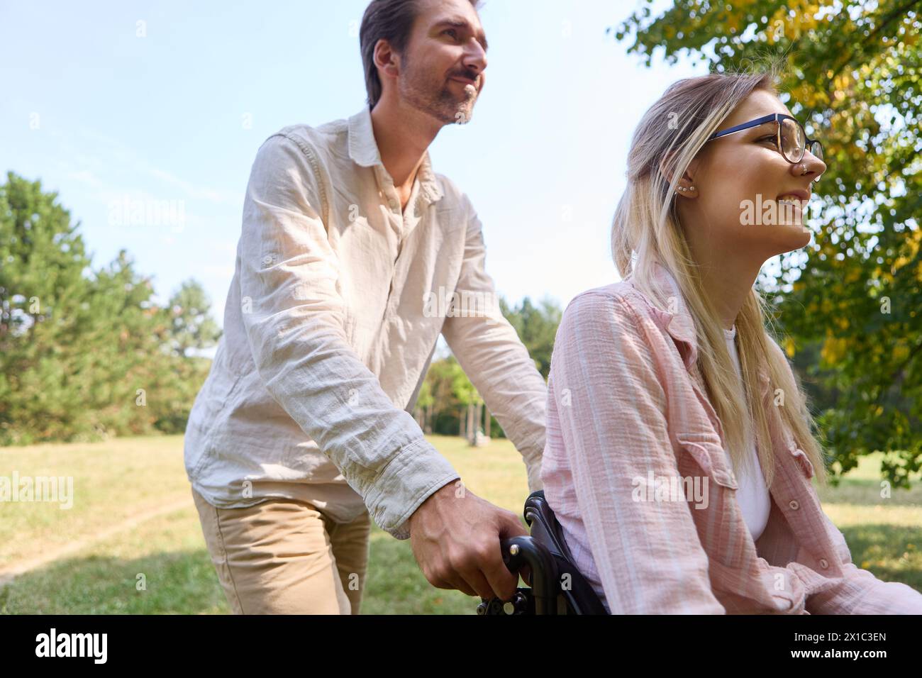 Ein Moment der Einbeziehung im Freien mit einer freudigen Frau im Rollstuhl, die von einem Mann in einer ruhigen Parklandschaft unterstützt wird. Stockfoto