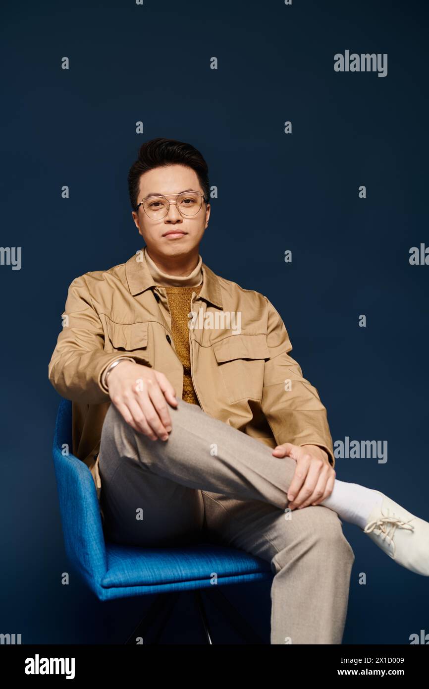 Ein modischer junger Mann in eleganter Kleidung sitzt auf einem blauen Stuhl und kreuzt seine Beine in selbstbewusster Pose. Stockfoto