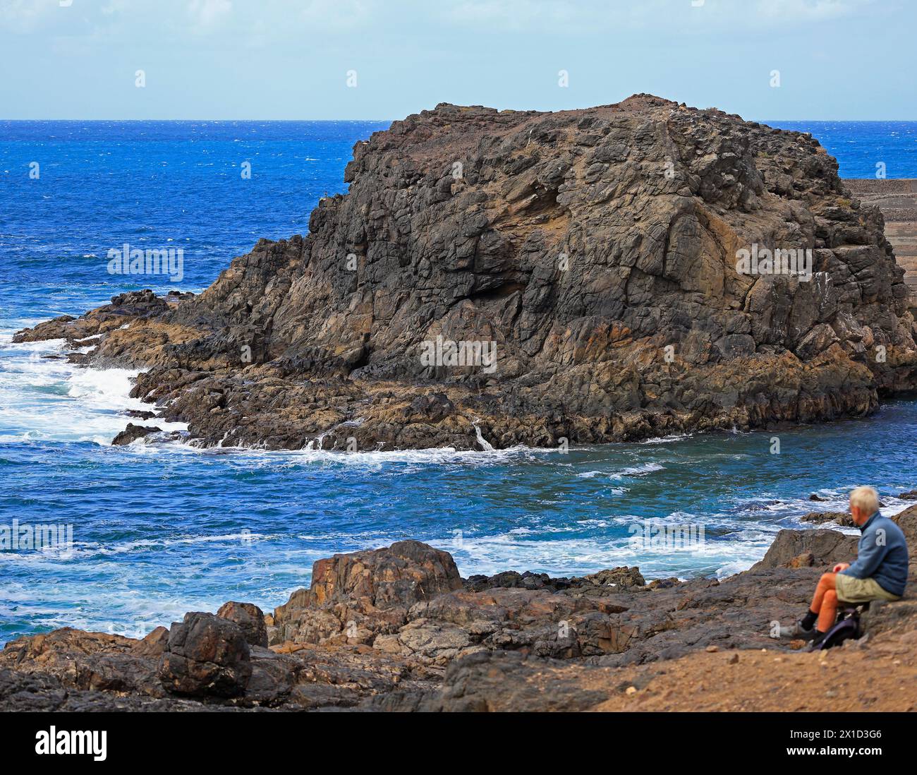 Mann sitzt und blickt auf die Aussicht, großer Felsvorsprung am Eingang zum Fischerhafen, El Cotillo, Fuerteventura, Kanarischen Inseln, Spanien. Vom Februar 2024 Stockfoto